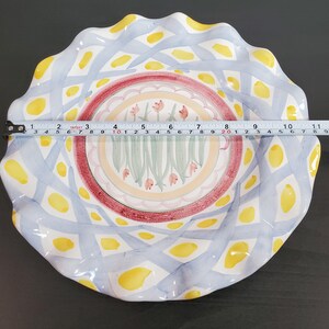 May include: A round ceramic plate with a scalloped edge, featuring a blue lattice pattern with yellow spots and a central floral motif of pink tulips. A measuring tape across the center shows a diameter of approximately 11 inches.