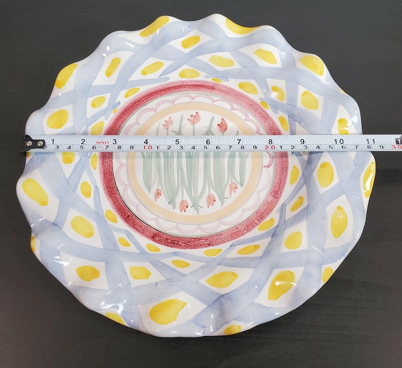 May include: A round ceramic plate with a scalloped edge, featuring a blue lattice pattern with yellow spots and a central floral motif of pink tulips. A measuring tape across the center shows a diameter of approximately 11 inches.