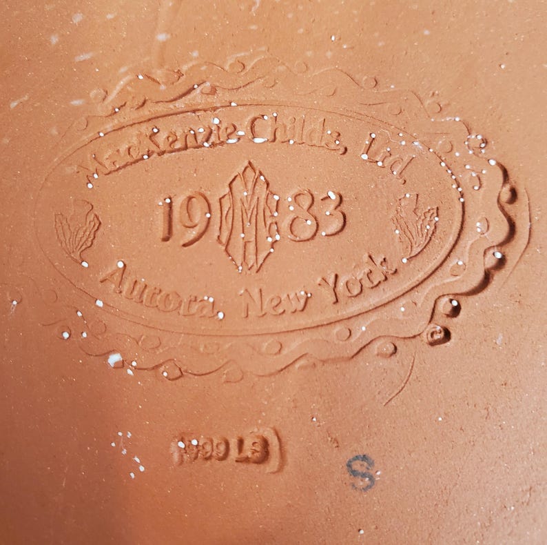 May include: A terracotta-colored ceramic surface featuring an embossed oval stamp with the text MacKenzie-Childs, Ltd. 1983 Aurora, New York. Below the oval, the text 1999 LS is embossed, and a small letter S is printed in black ink on the clay.