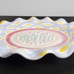 May include: A round ceramic serving platter with a ruffled, wavy edge. The rim features a light blue lattice pattern with yellow accents. The center displays a circular design with green leaf motifs and small pink flowers on a cream background.