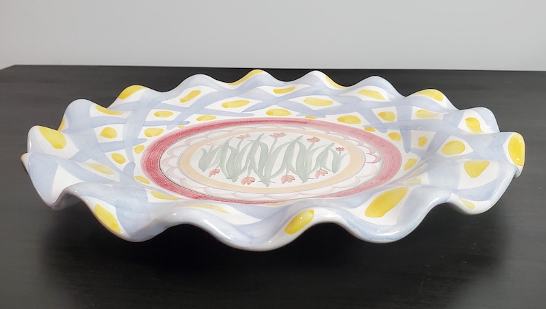 May include: A round ceramic serving platter with a ruffled, wavy edge. The rim features a light blue lattice pattern with yellow accents. The center displays a circular design with green leaf motifs and small pink flowers on a cream background.