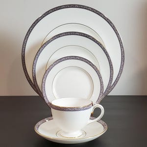 May include: A white porcelain tea cup and saucer set with a gold rim and a blue Greek key design. The set includes a variety of white plates with the same blue and gold design. Elegant tableware.