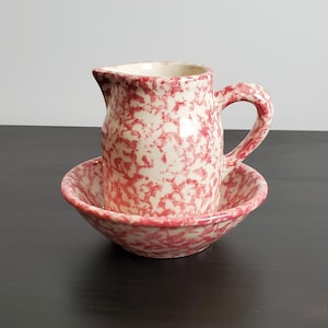 May include: A ceramic pitcher and matching bowl set featuring a cream-colored base with a speckled red pattern. The pitcher has a curved handle and a pouring spout, while the bowl has a rounded rim. Both pieces have a consistent mottled finish.
