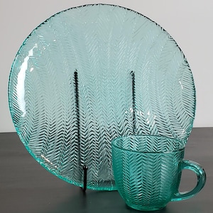 May include: A teal-colored glass plate and mug set. The plate is large and round, with a textured chevron pattern. The mug has a matching design and a handle. The set is displayed on a dark surface against a neutral background.