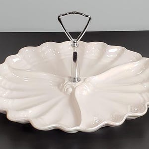 May include: A white ceramic serving tray with a scalloped edge and three divided sections. The tray features a central chrome-finished metal handle with a triangular loop at the top, allowing for easy transport of the dish.