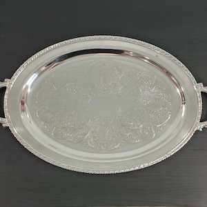 Puede incluir: Una bandeja de servicio ovalada de tono plateado con una superficie pulida y reflectante que presenta un intrincado patrón floral en relieve en el centro. La bandeja tiene un borde decorativo de cuerda y dos asas ornamentadas en los lados.