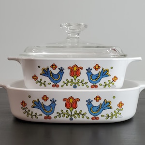 May include: Two white rectangular casserole dishes with integrated side handles, featuring a colorful pattern of blue birds, orange tulips, and green vines. The top dish includes a clear glass lid with a round knob handle. Both pieces are marked 1972.