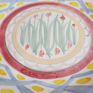 May include: A ceramic plate featuring a central circular motif of pink tulips with green stems and leaves. The center is surrounded by a scalloped pink border, a solid peach ring, and a wide textured red band. The outer rim has blue cross-hatched lines.