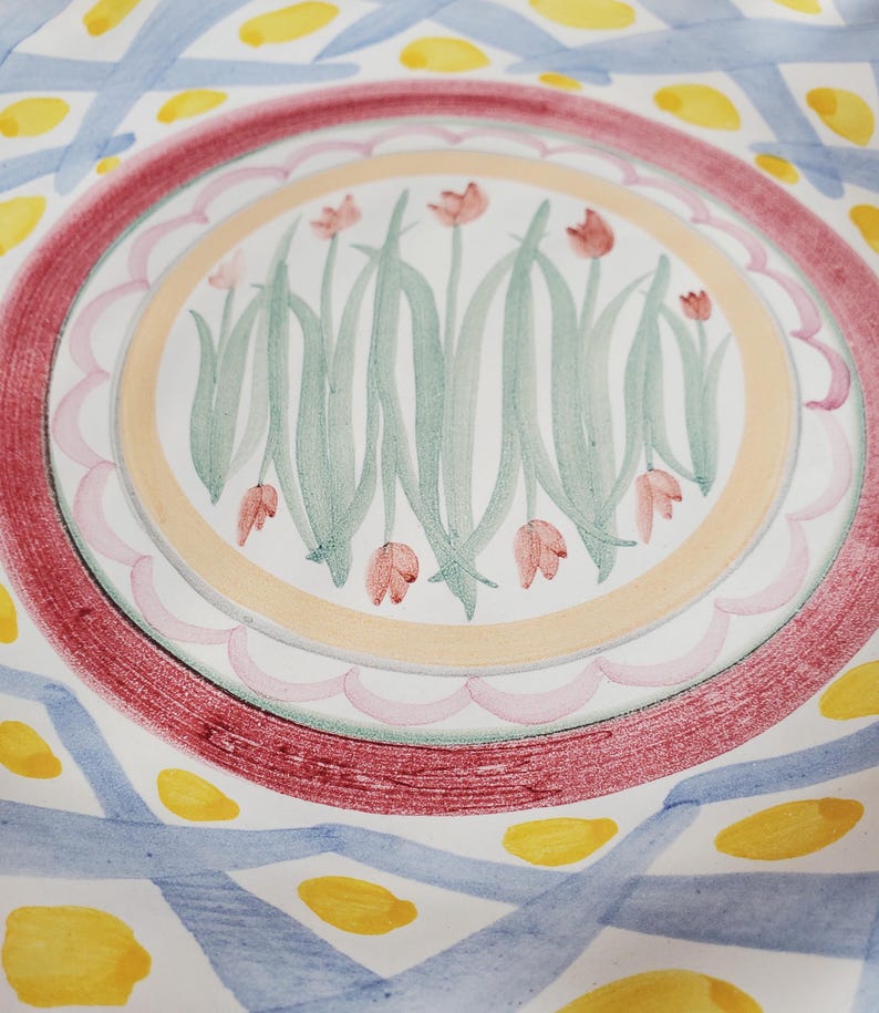 May include: A ceramic plate featuring a central circular motif of pink tulips with green stems and leaves. The center is surrounded by a scalloped pink border, a solid peach ring, and a wide textured red band. The outer rim has blue cross-hatched lines.