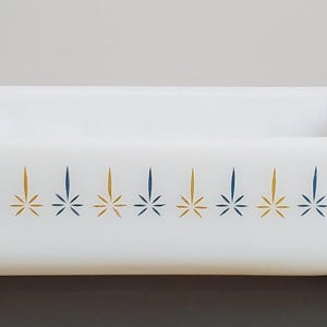 Puede incluir: Una fuente de horno rectangular de vidrio opalino blanco con asas laterales integradas. El exterior presenta un patrón horizontal repetitivo de motivos de estrellas azules y doradas alternadas en una sola fila central.