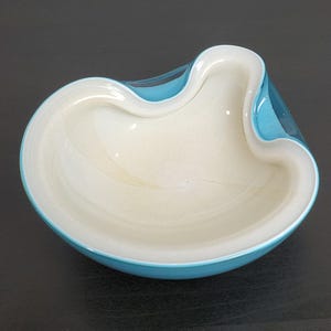 Puede incluir: Un cuenco de cristal con una forma orgánica y asimétrica, con un borde curvo y suave. El exterior del cuenco es de un azul turquesa vibrante, mientras que el interior es de un color blanco crema contrastante, creando un aspecto estratificado.
