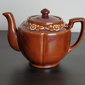 May include: A glossy brown ceramic teapot featuring a rounded body with vertical fluted panels and a matching lid. A decorative band of raised, cream-colored floral and leaf motifs encircles the upper shoulder of the teapot near the lid.