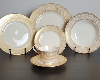 May include: A five-piece porcelain dinnerware set featuring a white base with intricate gold filigree patterns along the rims. The set includes a dinner plate, a salad plate, a soup bowl, and a teacup with a matching saucer, all with gold trim.