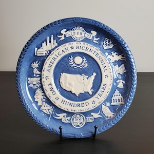Può includere: Un piatto commemorativo rotondo in ceramica blu e bianca con una mappa in rilievo degli Stati Uniti al centro. Il testo recita American Bicentennial Two Hundred Years 1776 1976, circondato da icone storiche e date.