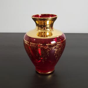 May include: A ruby red glass vase featuring a wide, polished gold-tone neck and rim. The body of the vase is decorated with an intricate, repeating gold filigree pattern that encircles the midsection, contrasting against the deep red glass.