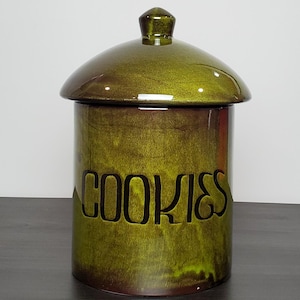 Può includere: Un barattolo per biscotti cilindrico in ceramica con una finitura lucida nei toni del verde oliva e del marrone screziato. Il barattolo presenta un coperchio rimovibile coordinato con un pomello centrale. La scritta COOKIES è in rilievo.