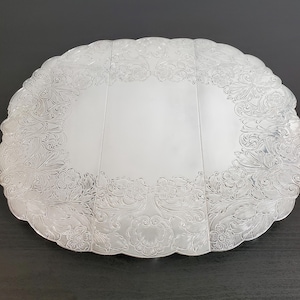 May include: A large, oval silver serving platter featuring a polished, smooth central area and a wide, scalloped border decorated with intricate, embossed floral and scrollwork patterns that extend across the entire outer edge of the tray.