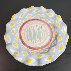 May include: A round ceramic plate with a ruffled edge featuring a central medallion of painted pink tulips and green stems. The border displays a lattice pattern of light blue brushstrokes interspersed with yellow spots on a white background.