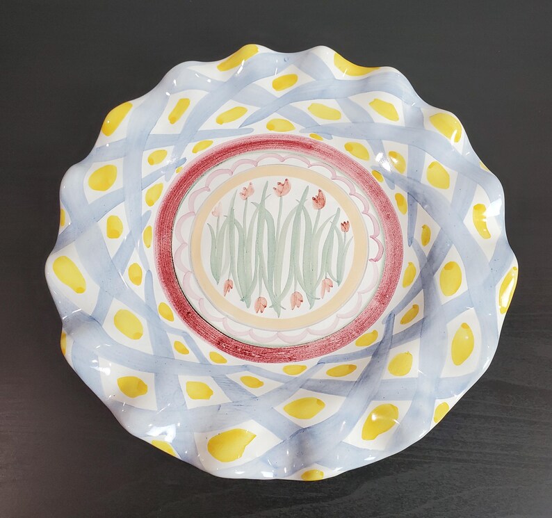May include: A round ceramic plate with a ruffled edge featuring a central medallion of painted pink tulips and green stems. The border displays a lattice pattern of light blue brushstrokes interspersed with yellow spots on a white background.