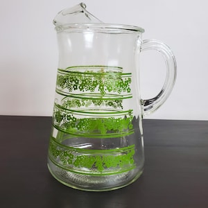 May include: A clear glass pitcher featuring a repeating pattern of horizontal green stripes and small floral motifs. The pitcher has a sturdy handle on the side and a wide pouring spout at the top rim, designed for serving beverages in a kitchen setting.
