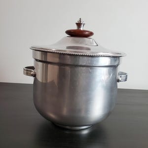 Pode incluir: Um balde de gelo de metal prateado com corpo arredondado, duas alças laterais e uma tampa removível. A tampa apresenta uma borda frisada e um botão de madeira circular castanho-escuro no topo. O balde tem um acabamento liso e reflexivo.