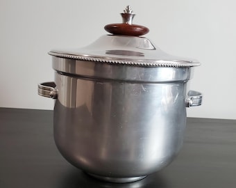 Puede incluir: Una cubitera de metal plateado con cuerpo redondeado, dos asas laterales y tapa extraíble. La tapa presenta un borde con cuentas y un pomo de madera circular de color marrón oscuro en la parte superior. El cubo tiene un acabado liso y reflectante.