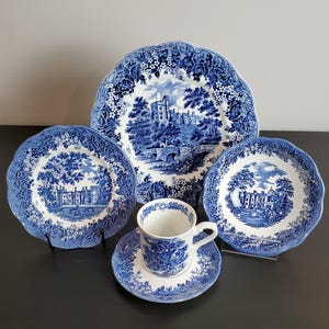 May include: A blue and white ceramic dinnerware set featuring a large platter, two smaller plates, and a teacup with saucer. Each piece displays detailed transferware illustrations of historic buildings, landscapes, and floral borders.