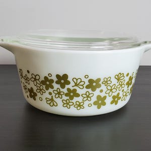 May include: A white glass casserole dish with a clear glass lid, featuring a repeating pattern of olive green flowers and small dots around the exterior. The dish has two integrated handles on opposite sides and a smooth, rounded base.