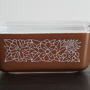 Può includere: Un piatto Pyrex rettangolare marrone con coperchio in vetro trasparente. Il piatto presenta un motivo floreale bianco sul davanti. Il piatto è progettato per la conservazione degli alimenti.