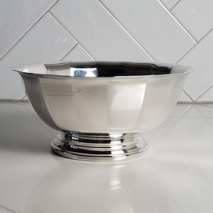 May include: A polished silver-toned bowl with a smooth, reflective surface and a faceted, multi-sided design. The bowl features a wide, open top that tapers down to a circular, tiered pedestal base, creating a classic, symmetrical silhouette.