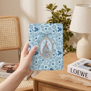 Puede incluir: Una funda azul claro con una ilustración de un castillo dentro de un marco ovalado, rodeada de un patrón floral y de Mickey Mouse. La funda está sujeta en la mano de una persona.