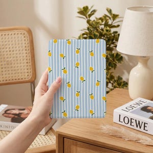 Puede incluir: Una funda rectangular para tableta con un patrón de rayas azules y blancas. La funda está decorada con limones amarillos y hojas verdes. La funda está siendo sostenida por una persona.