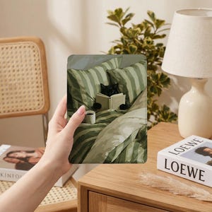May include: A rectangular e-reader case featuring a printed illustration of a black cat sitting in a bed with green and white striped pillows and blankets, reading an open book while a small white mug sits on the bed covers near the cat.