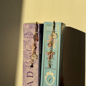 Può includere: Orecchini pendenti color oro con vari ciondoli, tra cui un fiocco rosa, un cuore, una busta e una stella. Gli orecchini sono esposti accanto a due libri intitolati "Mad Majesty" e "Inheritance". Design delicato e femminile.
