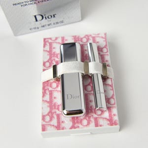 May include: A Dior ready-to-wear summer makeup set. The set includes two silver tubes secured with a white band on a pink and white patterned base. The box in the background has the Dior logo and text.
