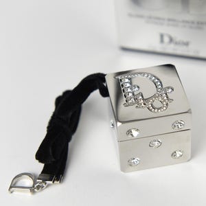 May include: A silver-coloured, square compact with a black velvet strap and a silver clasp. The compact features the Dior logo in rhinestones on the top and decorative rhinestones on the sides. A white box with the Dior logo is in the background.