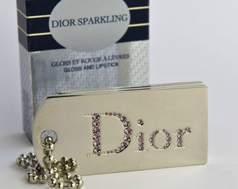 May include: A silver-toned rectangular Dior compact case featuring the brand name spelled out in small pink crystals. A silver ball chain is attached to the side of the case. Behind it stands a silver and navy box labeled DIOR SPARKLING 001.