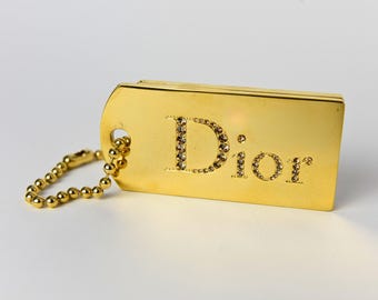 May include: A gold-colored rectangular tag with the word "Dior" spelled out in sparkling crystals. The tag is attached to a small gold ball chain. The tag has a hole at the top left corner.