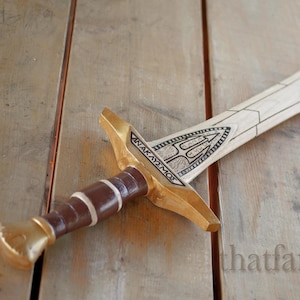 Op de afbeelding: Een houten zwaard met een gouden handvat en een bruine leren greep. Het blad is versierd met een drietandontwerp en de inscriptie "ANAKAYMOS".