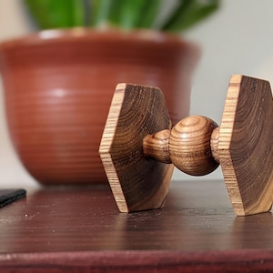 May include: A wooden Tie Fighter-shaped object with a central sphere and two angled, geometric wings. The wood grain is visible, and the object is set against a blurred background with a terracotta pot and a plant.