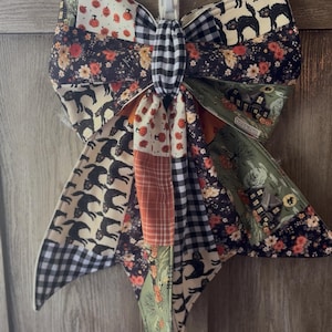 May include: A decorative fabric bow featuring a patchwork design with various patterns including black cats, orange pumpkins, floral prints, haunted houses, and black and white gingham checks. The bow is constructed from multiple fabric segments.