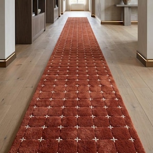 May include: Long, rectangular rust-colored rug with a grid pattern of white crosses. The rug is placed in a hallway with light wood flooring and a neutral-toned wall. The rug's design adds a touch of visual interest to the space.