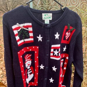 May include: Navy blue cardigan sweater with a festive holiday design. The sweater features red and white accents, including a snowman, stars, and gift boxes. The sweater has a button-up front and is perfect for the holiday season.