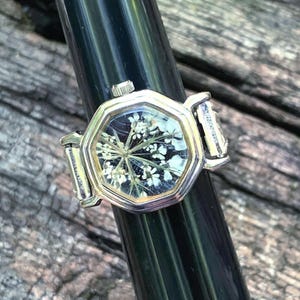 May include: A silver-toned ring with a watch face design. The octagonal face features a dark background with a delicate white floral arrangement. The ring is displayed on a black cylindrical object against a weathered wood surface.
