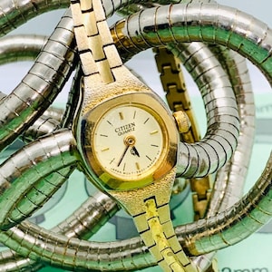 May include: A gold-toned Citizen Quartz watch with a cream-colored face and black hands. The watch is surrounded by a silver-colored, flexible metal band. The watch face is approximately 30mm in diameter.