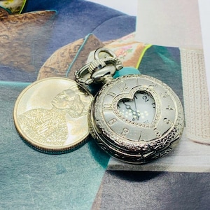 May include: Silver pocket watch with a heart-shaped window, next to a coin. The watch has a detailed design around the edges and a white face with black numerals. The watch is a quartz movement.