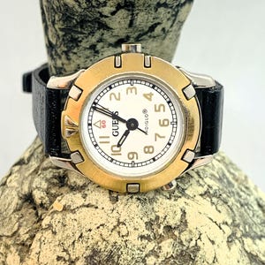 Può includere: Un orologio Guess color oro con quadrante bianco e cinturino in pelle nera. L'orologio ha un quadrante rotondo con indici orari in numeri romani neri e una lancetta dei secondi nera. Il quadrante dell'orologio presenta anche la scritta "Guess" stampata in lettere nere.
