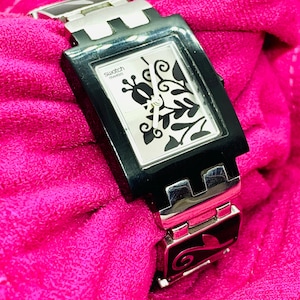May include: A rectangular Swatch watch with a black frame and silver band. The watch face is white with black floral designs. The watch is displayed on a bright pink fabric.