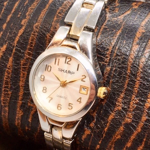 May include: A silver and gold tone women's wristwatch with a white face and black numbers. The watch face says "SHARP" and "QUARTZ".