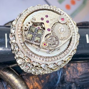 May include: A silver ring with a circular design featuring a watch movement with gears and a black stone. The ring is adorned with small, clear rhinestones.